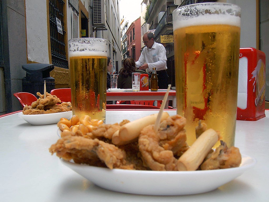 A waiter in the background of a table with two beers and some tapas